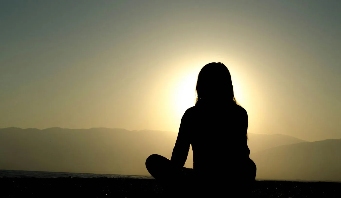 A woman sits cross-legged facing the setting sun